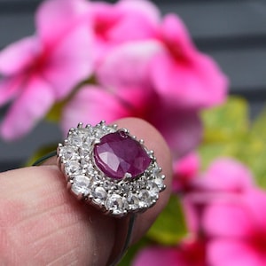 May include: A silver ring with a large, oval, deep red ruby gemstone, encircled by smaller, clear gemstones. The ring is set against a backdrop of blurred pink flowers, with a focus on the jewellery.