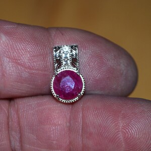 May include: A silver pendant featuring a large, round, faceted ruby gemstone. The ruby is a deep red colour. The pendant has an ornate, filigree design at the top, adding detail to the jewellery.