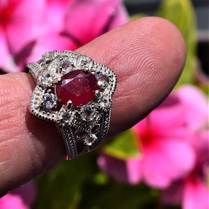 May include: A silver ring featuring a large, oval-cut ruby gemstone at its centre. The ring is adorned with smaller, clear gemstones and has an intricate, filigree pattern on the band. The ring is set against a backdrop of pink flowers.