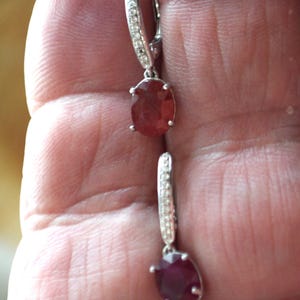 Peut inclure: Une paire de boucles d'oreilles en argent avec une pierre ovale rouge et une rangée de petites pierres claires sur le cercle.