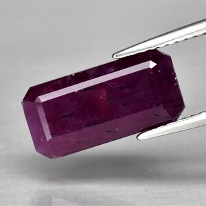 May include: A deep purple gemstone with a rectangular emerald cut. The gemstone is held by tweezers.