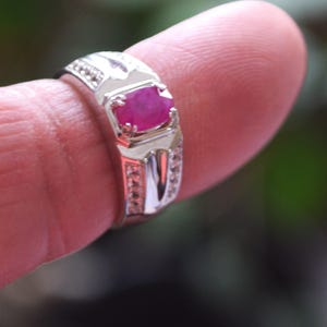 May include: A silver ring featuring a rectangular, vivid red gemstone. The band is embellished with smaller, clear stones and decorative elements. The ring is displayed on a finger, with a blurred green backdrop.