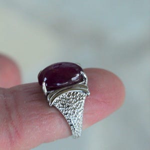 May include: A silver ring with a large, oval, deep red gemstone. The ring's band has a textured, studded design, and the gemstone is set in a smooth, polished bezel. The ring is a piece of jewellery.