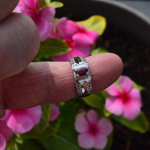 May include: A silver ring featuring a rectangular, deep red gemstone. The ring has a decorative design with smaller, clear stones and white accents. The ring is displayed against a backdrop of pink flowers and green foliage.
