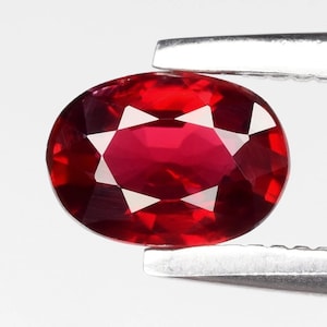 Könnte beinhalten: Ein facettierter, ovalförmiger Rubin-Edelstein, der von einer silbernen Pinzette gehalten wird. Der Rubin ist ein tiefes, leuchtendes Rot mit mehreren Facetten, die das Licht reflektieren. Der Edelstein ist ein Blickfang.