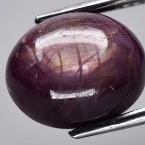 May include: A round, deep purple gemstone with a cabochon cut. The gemstone has a reflective surface and a slight shimmer.