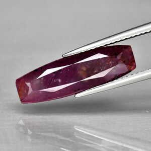 May include: A deep red ruby gemstone with a faceted, rectangular shape. The ruby is held in a pair of tweezers.
