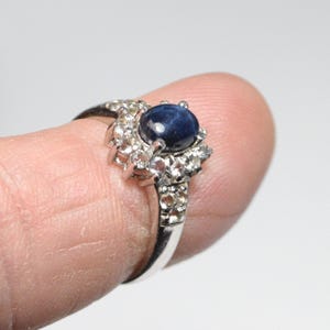 Könnte beinhalten: Ein silberner Ring mit einem großen, runden, dunkelblauen Edelstein in der Mitte, umgeben von kleineren, klaren Steinen. Der Ring wird von einem Finger vor weißem Hintergrund gehalten. Der Ring hat ein florales Design.