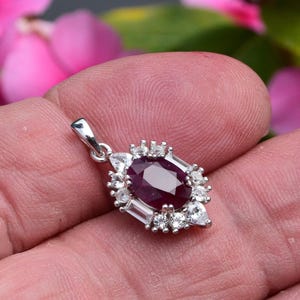 May include: A silver pendant with a central oval ruby, encircled by smaller, clear gemstones. The ruby is a rich red hue, with the surrounding stones in round and baguette cuts. The pendant is held in a hand, with a blurred floral backdrop.