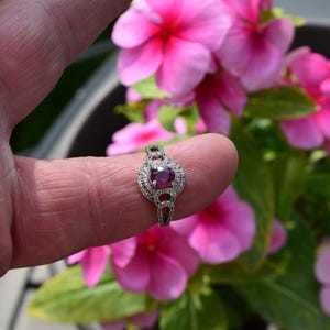 May include: A silver ring featuring an oval ruby gemstone, encircled by smaller clear stones. The ring is displayed against a background of bright pink flowers and green leaves.