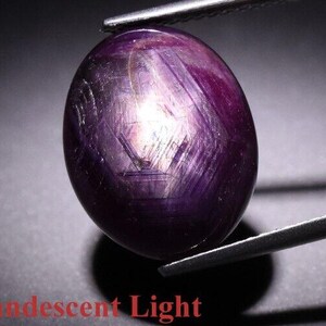 May include: A close-up of a deep purple oval-shaped gemstone held by tweezers. The gemstone has a star-like pattern and is illuminated by incandescent light.