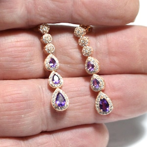 May include: A pair of gold-coloured drop earrings. Each earring has a series of small, round, clear stones leading to three pear-shaped, deep purple gemstones. The purple stones are surrounded by a halo of smaller, clear stones.