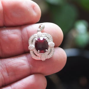 May include: A silver pendant featuring a large, oval, deep red gemstone. The gemstone is encircled by a halo of smaller, clear stones in a floral design. The pendant has a small loop at the top for a chain, suitable for necklaces.