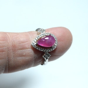 May include: A silver ring with a striking, pear-shaped pink gemstone, encircled by a halo of small, clear stones. The ring's band is also embellished with smaller stones, producing a sparkling appearance. The ring is a piece of jewellery.