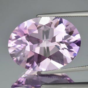 May include: A faceted, oval gemstone in shades of lavender and light purple. The gem is held by tweezers, highlighting its intricate cut and reflective surfaces. The gemstone's facets create a play of light and shadow.