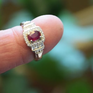 Könnte beinhalten: Ein goldener Ring mit einem rechteckigen, rubinroten Mittelstein, umgeben von kleineren, klaren Steinen. Der Ring wird vor einem verschwommenen grünen Hintergrund präsentiert, der das Design und den Glanz des Schmuckstücks hervorhebt.