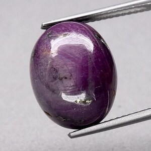 May include: A deep purple oval-shaped gemstone with a high polish. The gemstone has a slightly cloudy appearance with flecks of gold.