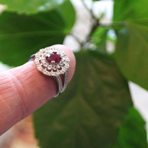 May include: A silver ring featuring a red ruby gemstone encircled by smaller white gemstones. The ring has a delicate, floral design.