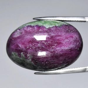 Peut inclure: Une pierre précieuse ovale polie, tenue par une pince. La pierre présente un dégradé de couleurs saisissant, passant d'un magenta profond au centre à du vert en haut et une teinte plus foncée à la base. La surface réfléchit la lumière.
