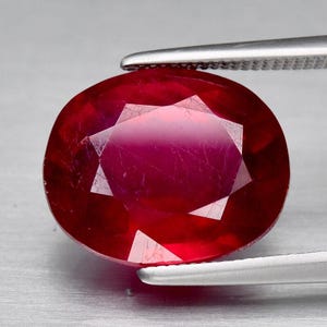 Peut inclure: Une pierre précieuse de forme ovale, un rubis facetté, maintenu par une pince argentée. La gemme est d'un rouge profond et saturé, avec des inclusions internes et une surface brillante et réfléchissante.