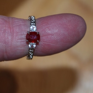 May include: A silver ring featuring a red oval gemstone and two smaller clear gemstones. The ring has a delicate, intricate design.