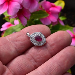 May include: A silver pendant featuring an oval ruby gemstone, encircled by smaller clear stones. The pendant is held in a hand, with pink flowers visible in the background.