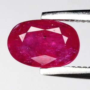 May include: An oval-cut ruby gemstone, held by silver tweezers. The ruby is a deep, saturated red with internal facets that reflect light. The gemstone is set against a white background.