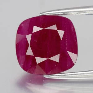 May include: A cushion-cut ruby gemstone, held by tweezers. The ruby is a deep red colour with facets that reflect light. The gemstone has a square shape with rounded corners and a star-like pattern in the centre.