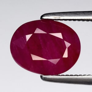 May include: Oval-cut ruby gemstone held by tweezers. The ruby is a deep, rich red colour with internal facets creating a starburst pattern. The gemstone is polished and has a smooth surface, reflecting light. The tweezers are silver.