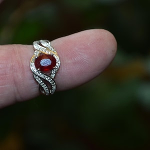 May include: A silver ring with a large, oval, red gemstone at the centre. The ring has a swirling design with small, clear accent stones. The ring is displayed on a finger against a blurred green background.