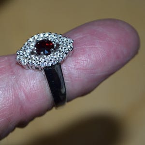 May include: A silver ring featuring a central dark red gemstone, encircled by smaller, clear stones. The ring is displayed on a finger, highlighting the detailed design and sparkle of the jewellery.