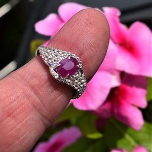 Peut inclure: Une bague en argent ornée d'un grand rubis ovale au centre, entouré de petits diamants transparents. La bague est présentée sur un fond de fleurs roses et de feuillage vert.