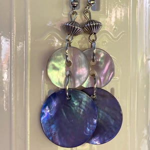 Può includere: Un paio di orecchini pendenti con dischi rotondi iridescenti in madreperla nei toni del viola, rosa e verde. Gli orecchini hanno dettagli in metallo argentato e ganci per le orecchie. Il design include piccole perline e un elemento a forma di cono a più livelli.