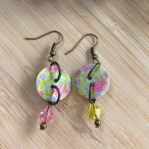 Könnte beinhalten: Blumenohrringe im Vintage-Stil. Jeder Ohrring hat eine runde, gemusterte Scheibe in Grün, Rosa und Lila mit einer daran hängenden rosa oder gelben Kristallperle. Die Ohrringe haben bronzefarbene Ohrhaken.