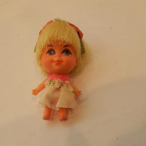 May include: A small vintage doll with blonde hair and blue eyes. The doll wears a white dress with a pink collar and floral accents. A pink bow adorns the doll's hair. The doll is a collectible item.