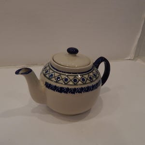 May include: Cream-colored ceramic teapot with a blue handle, spout, and lid. The teapot features a blue and green geometric pattern around the body. The lid has a small blue knob. A classic design for serving tea.