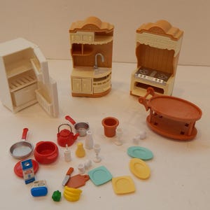 May include: A collection of miniature dollhouse kitchen furniture and accessories. Includes a white refrigerator, two tan cabinets, a stove, a table, and various red, white, and blue kitchenware items. The set is designed for play.