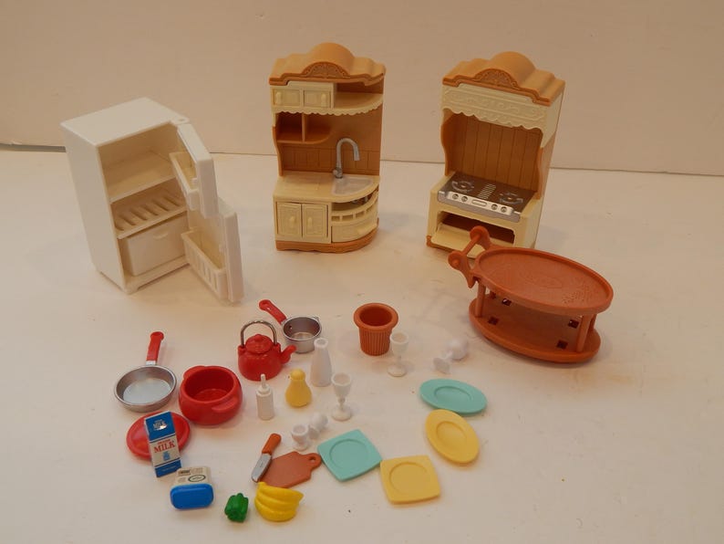 May include: A collection of miniature dollhouse kitchen furniture and accessories. Includes a white refrigerator, two tan cabinets, a stove, a table, and various red, white, and blue kitchenware items. The set is designed for play.