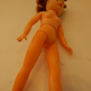 May include: A plastic doll with light peach-colored skin, red painted fingernails, and reddish-brown hair. The doll has a molded torso, articulated arms and legs, and facial features including red lips and dark eyes, shown from a high angle.
