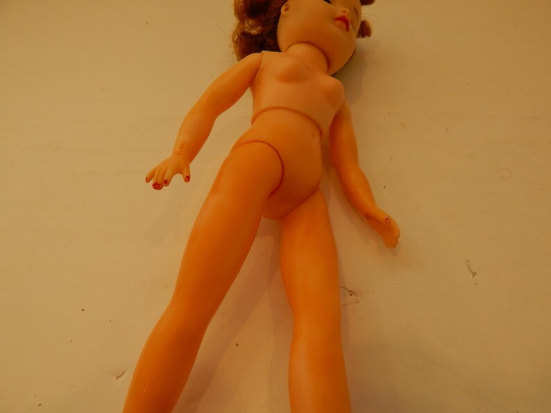 May include: A plastic doll with light peach-colored skin, red painted fingernails, and reddish-brown hair. The doll has a molded torso, articulated arms and legs, and facial features including red lips and dark eyes, shown from a high angle.