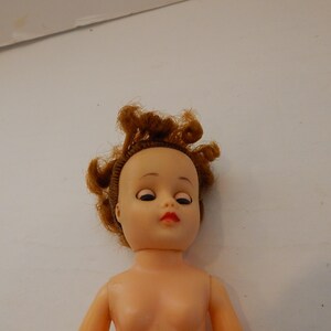 May include: A doll with a light peach-colored plastic head and torso. The doll features rooted, curly auburn hair, painted eyebrows, dark eyelashes, and bright red lips. The eyes are positioned to look downward, and the head is attached to a neck joint.
