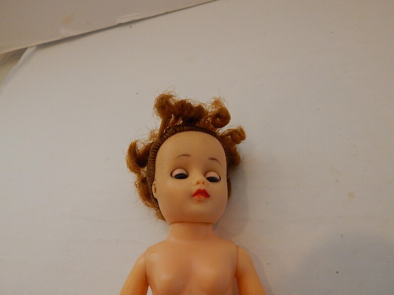 May include: A doll with a light peach-colored plastic head and torso. The doll features rooted, curly auburn hair, painted eyebrows, dark eyelashes, and bright red lips. The eyes are positioned to look downward, and the head is attached to a neck joint.