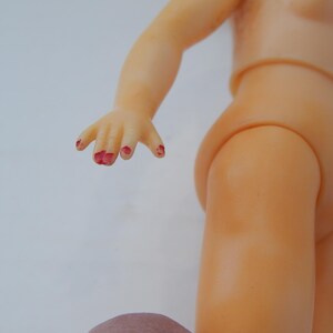 May include: A light-skinned plastic doll torso and arm with a hand featuring chipped red paint on the fingernails. The doll has a smooth, molded surface and a jointed shoulder connection, showing a partial view of the upper body and arm.