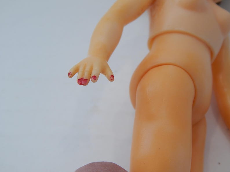May include: A light-skinned plastic doll torso and arm with a hand featuring chipped red paint on the fingernails. The doll has a smooth, molded surface and a jointed shoulder connection, showing a partial view of the upper body and arm.