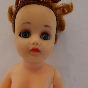 May include: A doll with a pale skin tone, light blue eyes, and red lips. The doll has rooted, curly reddish-brown hair styled in a partial updo. The facial features include painted eyebrows and eyelashes, with a smooth, molded plastic head and torso.