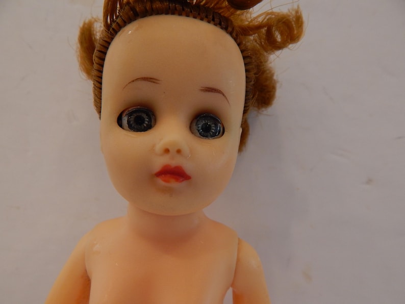 May include: A doll with a pale skin tone, light blue eyes, and red lips. The doll has rooted, curly reddish-brown hair styled in a partial updo. The facial features include painted eyebrows and eyelashes, with a smooth, molded plastic head and torso.