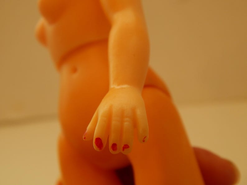May include: A peach-colored plastic doll torso and arm featuring a hand with red painted fingernails. The doll has a smooth, matte finish and a molded navel. The arm is positioned downward, showcasing the detailed fingers with red paint.