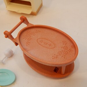 May include: An orange plastic serving tray with a handle, oval top, and floral design. The tray is part of a miniature toy set, with a small plate and wine glass visible nearby. The tray is approximately 4 inches long.