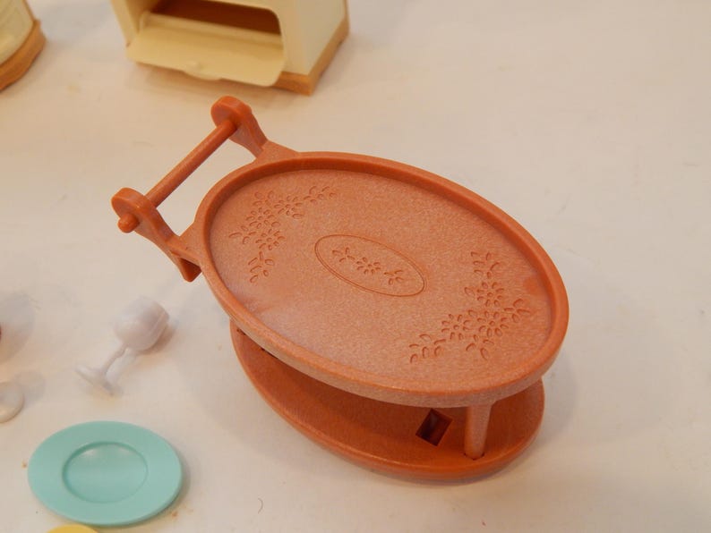 May include: An orange plastic serving tray with a handle, oval top, and floral design. The tray is part of a miniature toy set, with a small plate and wine glass visible nearby. The tray is approximately 4 inches long.