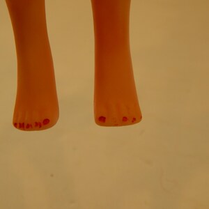 May include: A pair of light peach-colored doll feet with five toes on each foot. The toes are painted with a bright red color, resembling nail polish. The feet are positioned vertically, showing the front view of the toes and the lower legs.